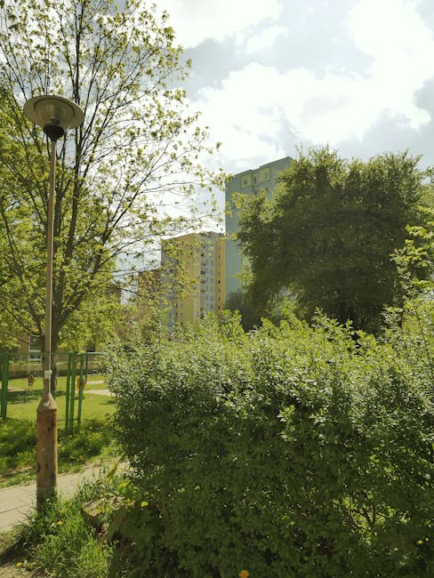 A small brown bird with a pale eye and a white wing bar is perched on a weathered piece of wood within a lush garden scene. Surrounding the bird are tall, green leafy plants with broad, variegated lea